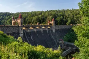 Malebný výhled na historickou přehradu s věžemi s červenými střechami, obklopenou bujnou zelení pod jasnou modrou oblohou.